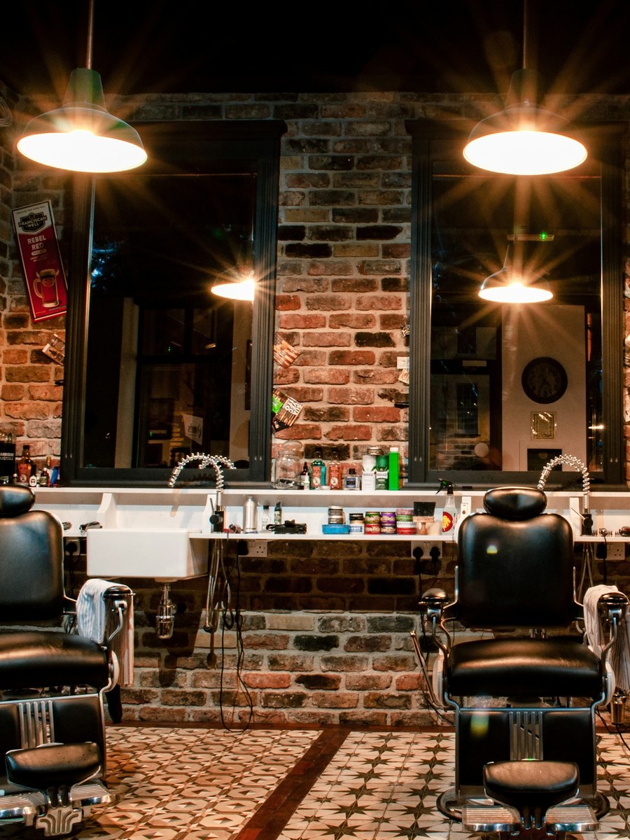 A barber shop under low pendant lamps, two chairs facing a run of mirrors set into a bare brick wall
